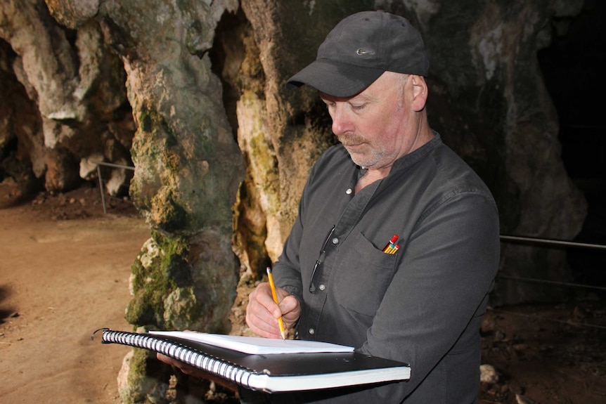 Julian Hume stands in a cave sketching on a notepad. Wearing black shirt with pencils tucked in front pocket and a black hat.