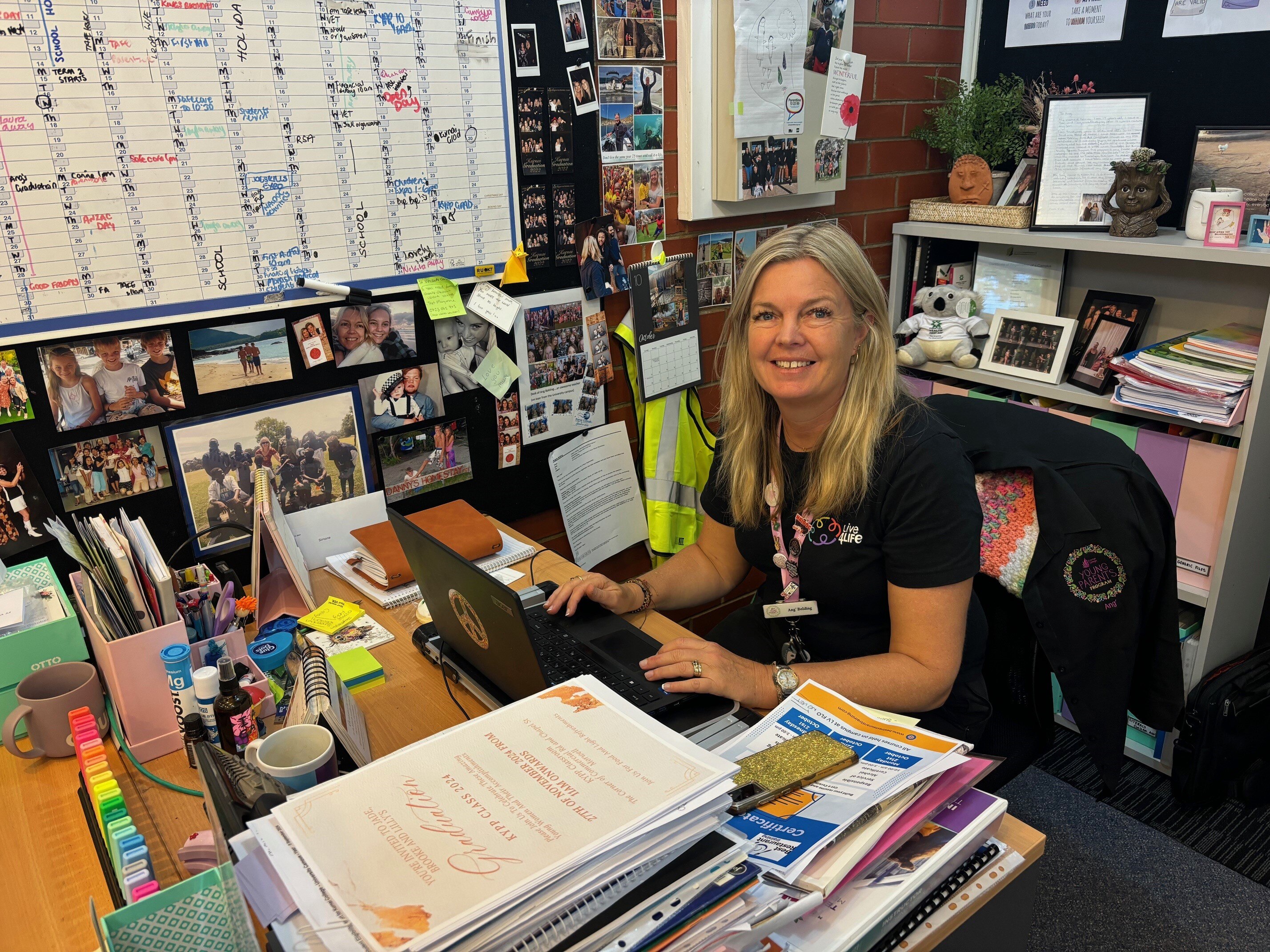 A smiling blonde woman, wears black, lanyard around neck, works on laptop on desk, in office full of pics, whiteboard.