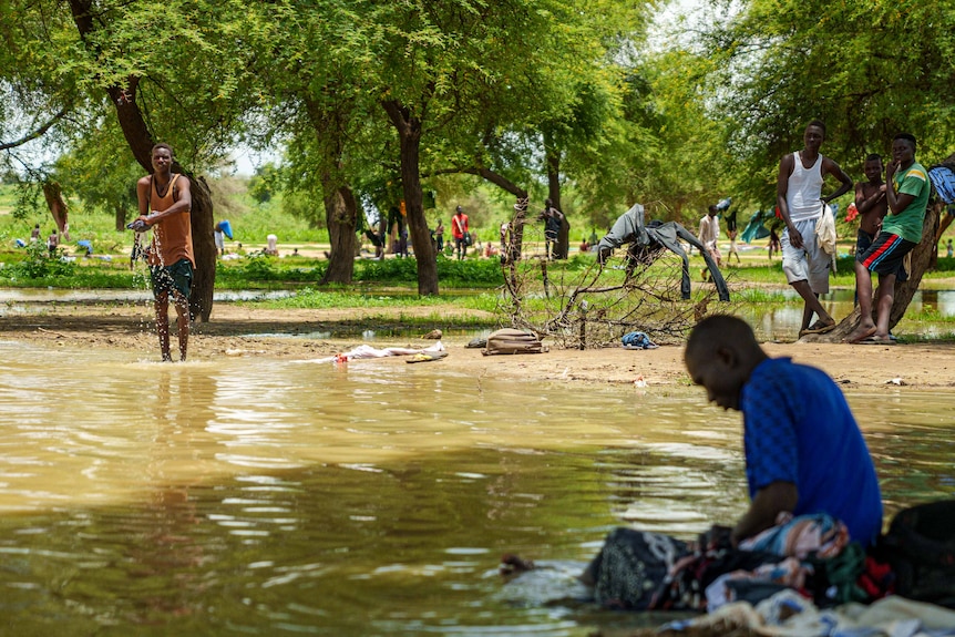 A man stands in a river in Chad surrouned by other men cooling down in the shade of a tree.