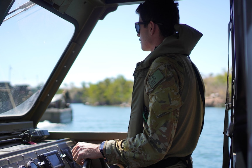 A man wearing sunglasses and a khaki jacket looks out onto the water as he drives a boat.