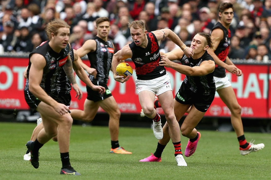 An Essendon AFL player holds the ball with his right hand as he is tackled by a Collingwood opponent.