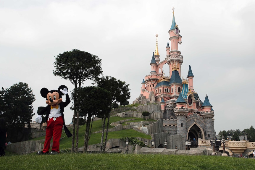 A person dressed as Mickey Mouse poses in front of the castle at Disneyland Paris.