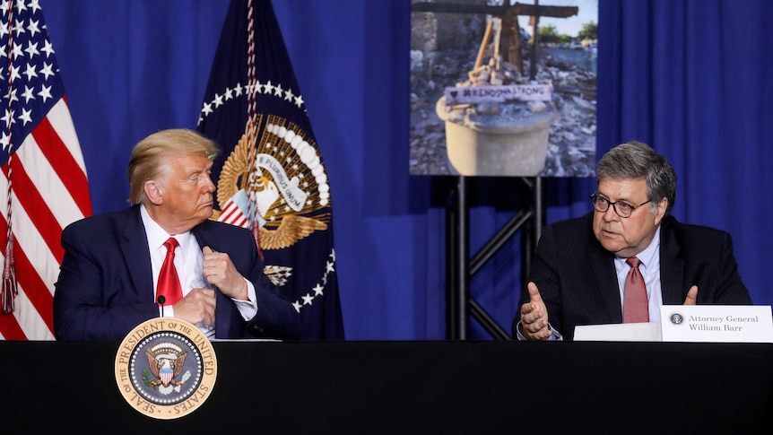 You see two men in suits sitting at a desk in front of an american flag and a photo of rubble from protests.