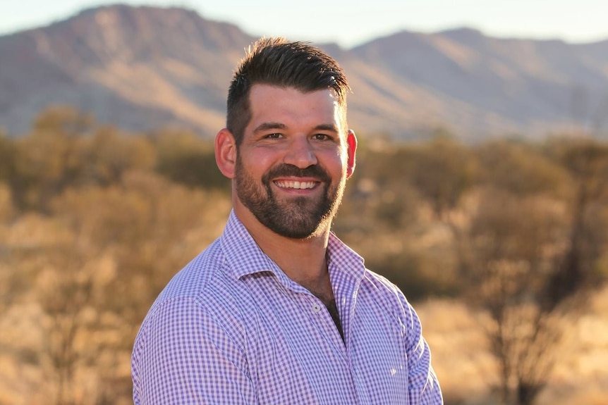 A bearded man in a checked shirt stands in front of a rocky mountain range.