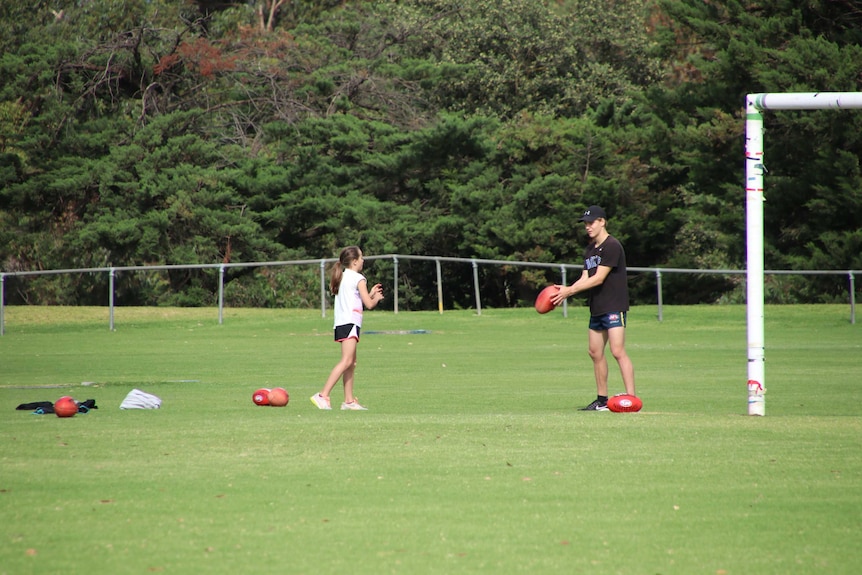 A young girl and teenage boy stand apart on an oval passing footballs on a sunny day.