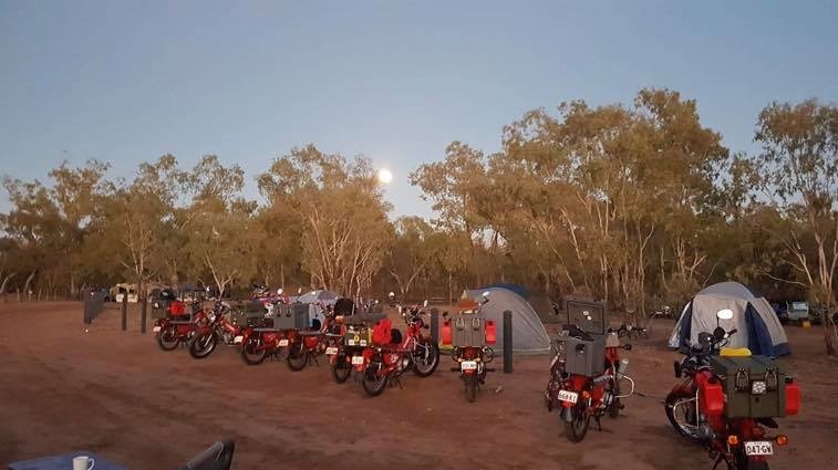 Red and black motorbikes with large cases are parked on the dirt next to a camping ground with tents pitched under trees.