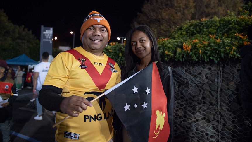 A man in a yellow and red jersey with a flag smiles next to a younger woman