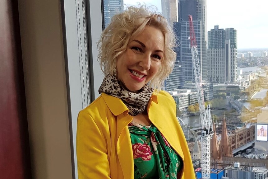A woman with blonde hair, wearing a floral blouse and yellow jacket, smiles for a posed photo.