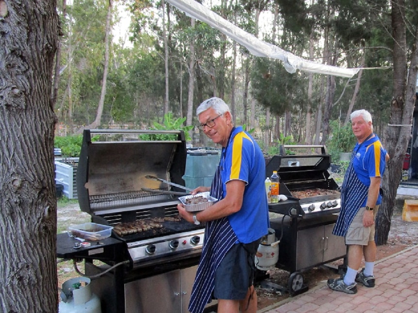 Two men stand at barbeques cooking food and smiling at the camera.