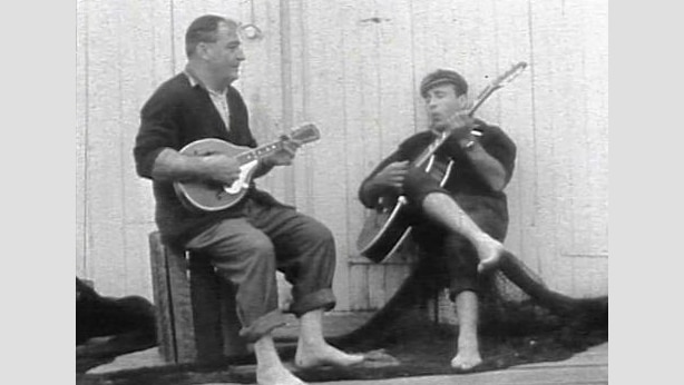 Two men sit on wooden crates while playing guitars