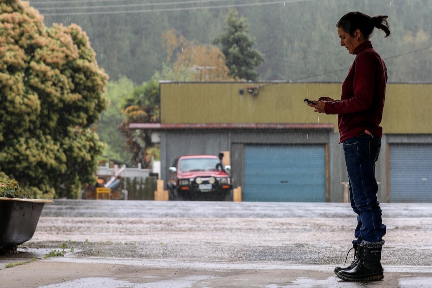 Woman in red jumper and blue jeans with greying black hair stands undercover with rain falling against tree-lined background