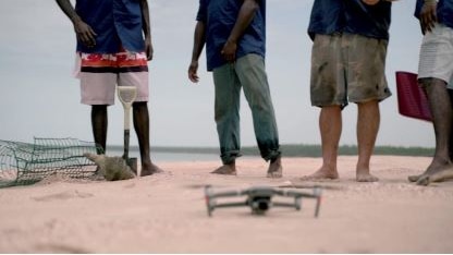 Four men stand on the sand in front a drone, a shovel and some protective netting used to