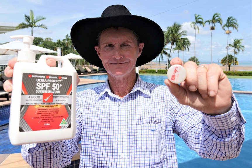 Billy Lowery wearing a cowboy hat holding lip balm and suncream to the camera beside a swimming pool
