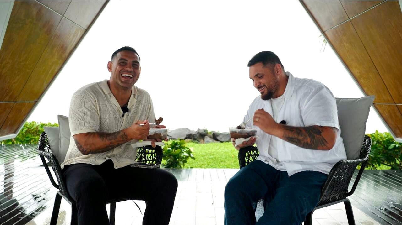 Two Samoan men wearing white shirts, dark pants & lavalava.  One appears to be laughing looking at camera while the other eats
