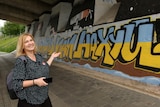 A woman with shoulder-length blonde hair, wearing a blouse and carrying a backpack, stands under a bridge, looking at graffiti