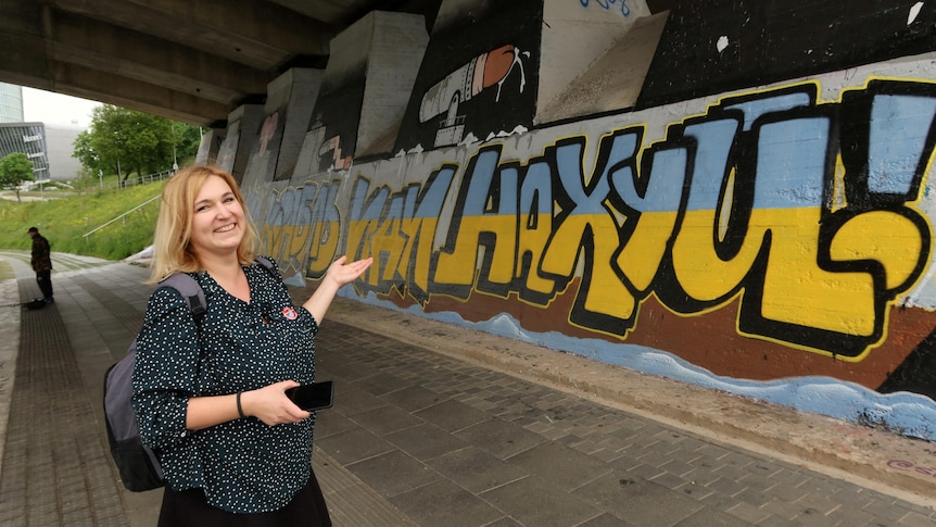 A woman with shoulder-length blonde hair, wearing a blouse and carrying a backpack, stands under a bridge, looking at graffiti