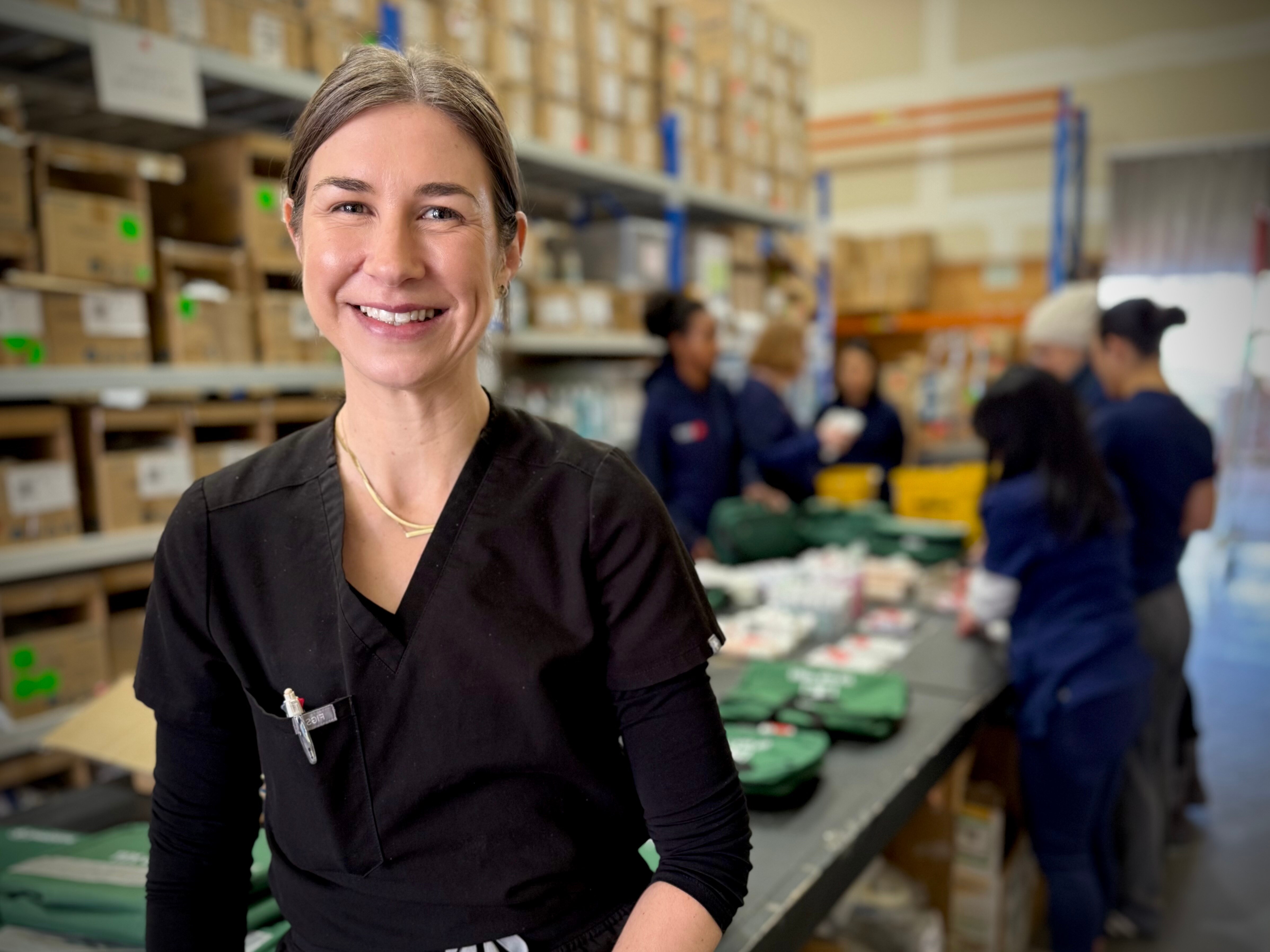 Uma mulher de cabelos e olhos castanhos está em um armazém cheio de suprimentos médicos. 