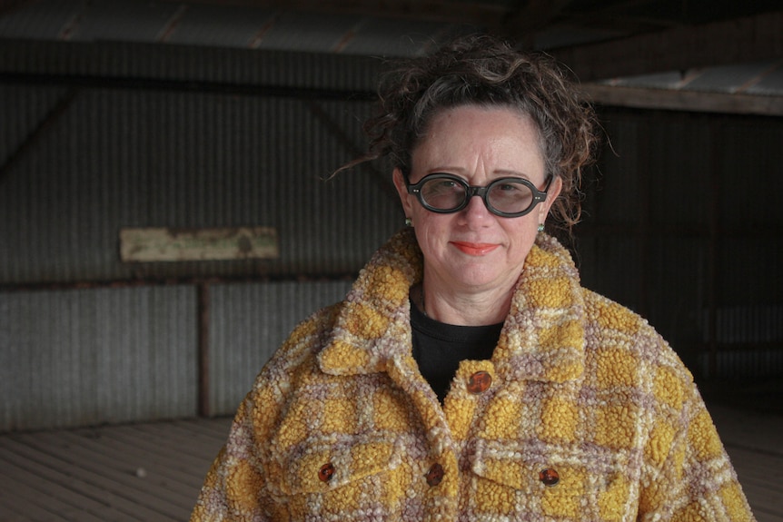 A woman wearing glasses and a yellow checked coat in a shed slightly smiling at the camera.