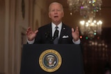 US President Joe Biden speaking at a lecturn with the presidential seal on it, wearing a suit and gesturing
