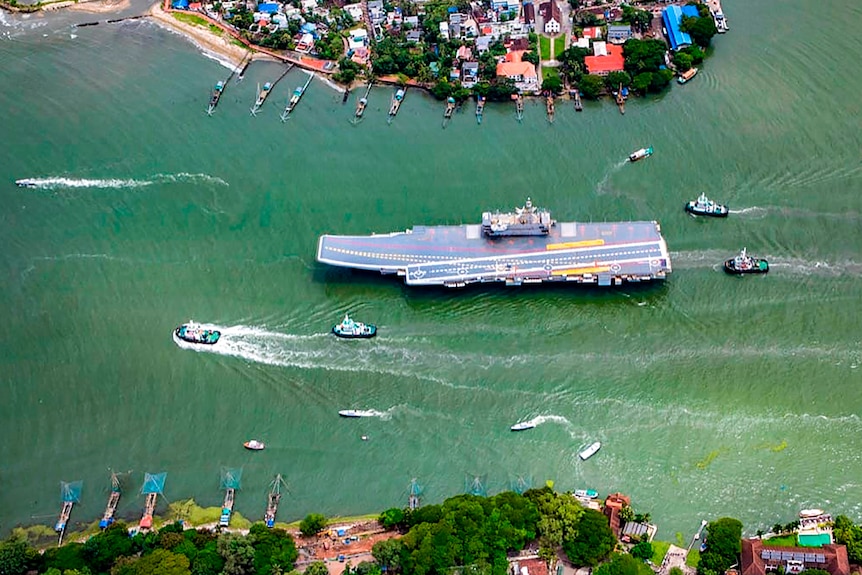 An aerial shot shows a large aircraft carrier being escorted by smaller vessels through a narrow pass.