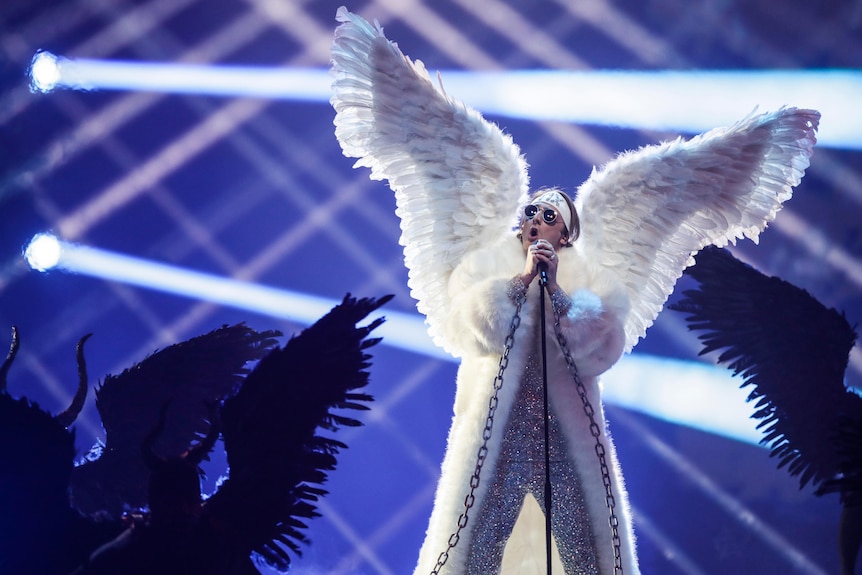 A man wearing huge white angel wings stands on stage and sings at the Eurovision Song Contest.