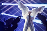 A man wearing huge white angel wings stands on stage and sings at the Eurovision Song Contest.