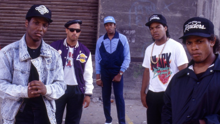 The members of N.W.A. stand around on the street wearing tracksuits and baseball caps