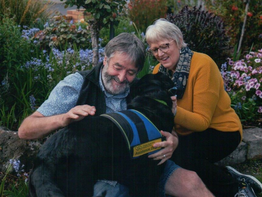 A man and a woman outside in a backyard garden hugging a black assistance dog