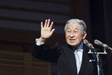 Japanese Emperor Akihito waves goodbye to well-wishers wearing a black suit with a silver tie in front of microphones.