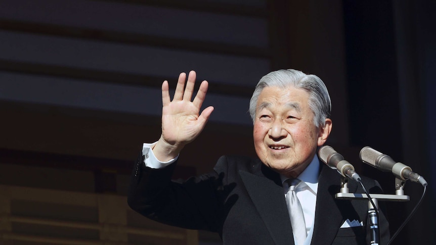 Japanese Emperor Akihito waves goodbye to well-wishers wearing a black suit with a silver tie in front of microphones.