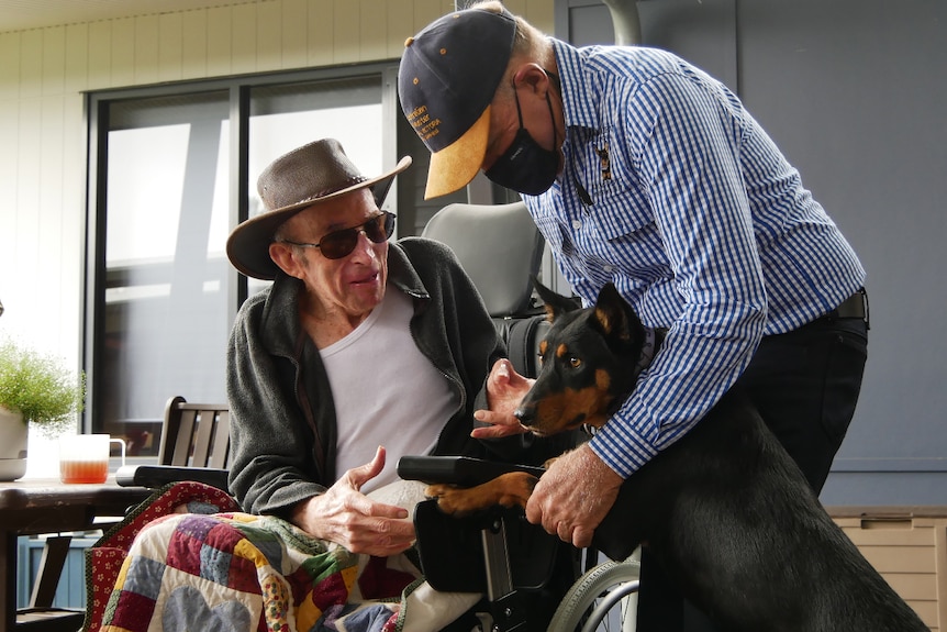 Frank Finger leaning over holding Annie's paws up on a man's chair, older man wearing hat and sunglasses reaching for dog.
