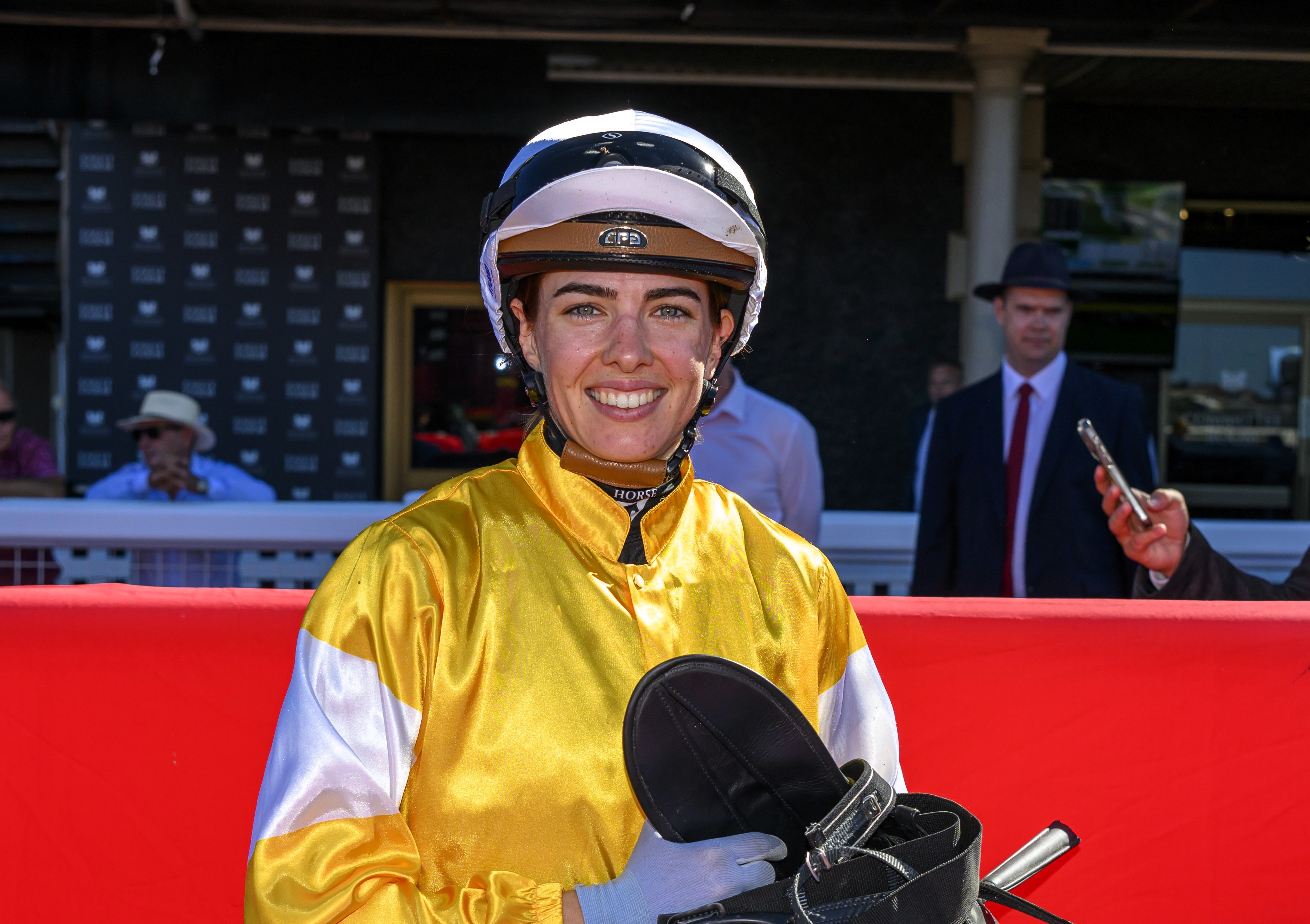 Um jóquei vestido de seda amarela e branca sorrindo com a sela na mão.