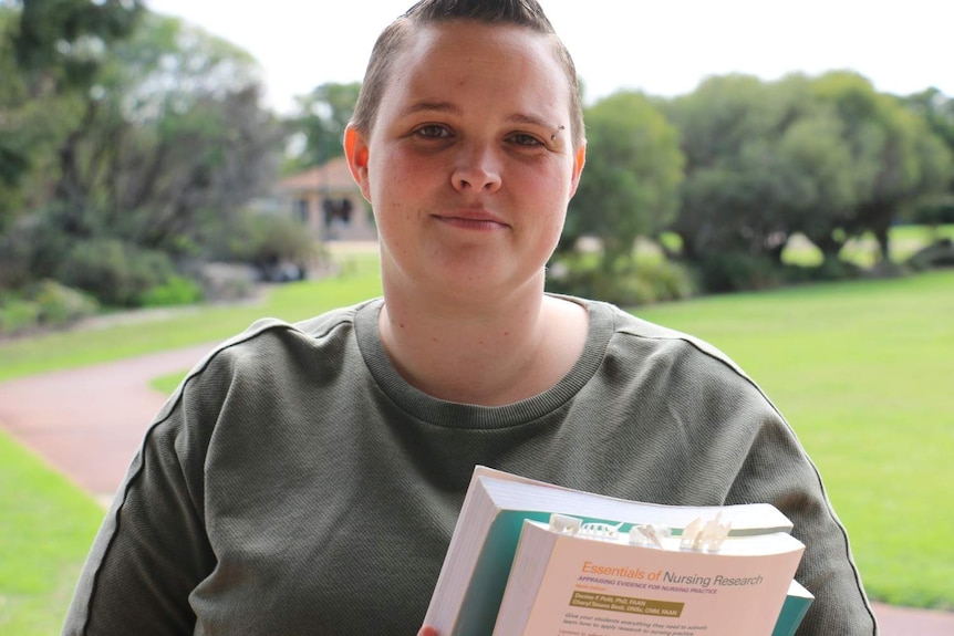 A woman wearing a sweater and carrying books smiles at a camera