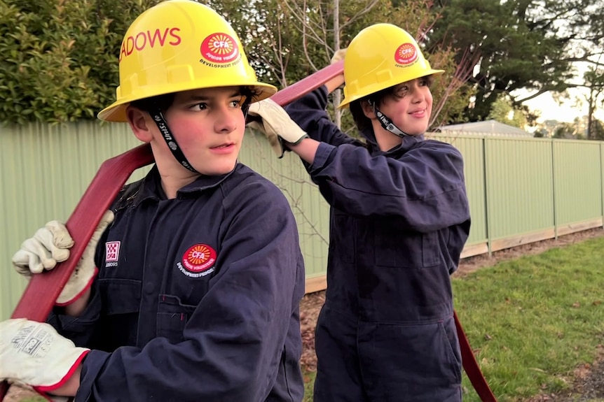 A boy and a girl wearing yellow hard hats and navy overalls with a red hose over their shoulder. 