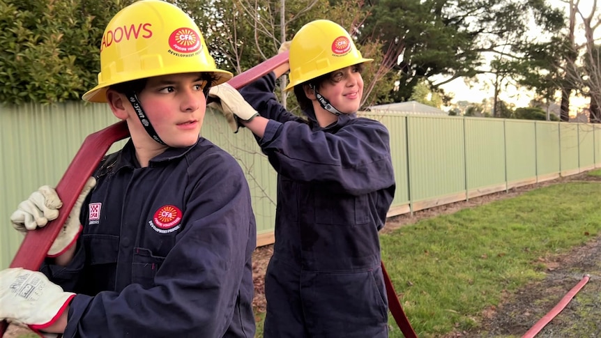 A boy and a girl wearing yellow hard hats and navy overalls with a red hose over their shoulder. 