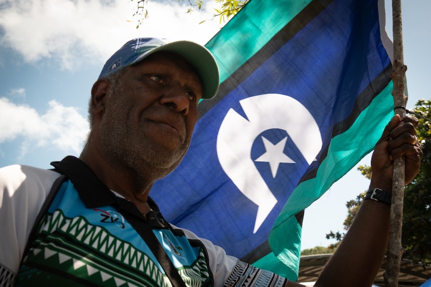 An indigenous man waves a blue, aqua and black striped flag with a 5-pointed white star inside a curved headdress design
