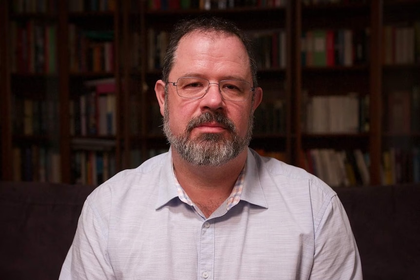 A man with a button-up shirt, glasses and beard stares at the camera.