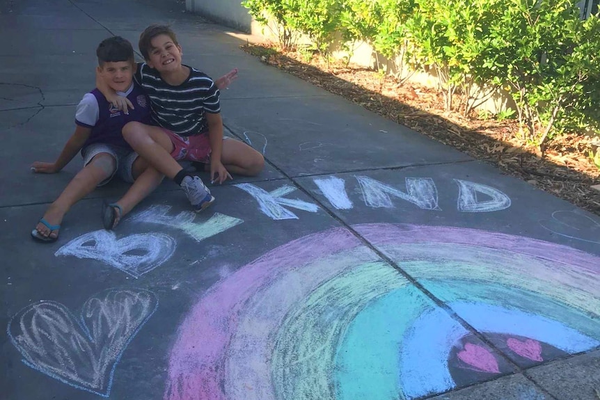 Two boys with their arms around each other sit in front of a chalk drawing saying be kind.