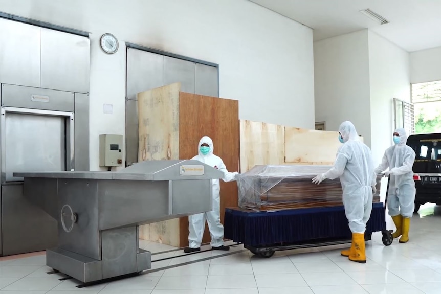 The inside of a crematorium where two men in protective clothing are standing in front of coffins. 
