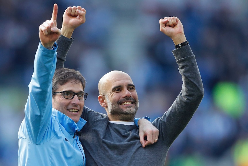 Pep Guardiola stands with his hands aloft wearing a grey cardigan, a member of his staff to his right drapes his arm around him