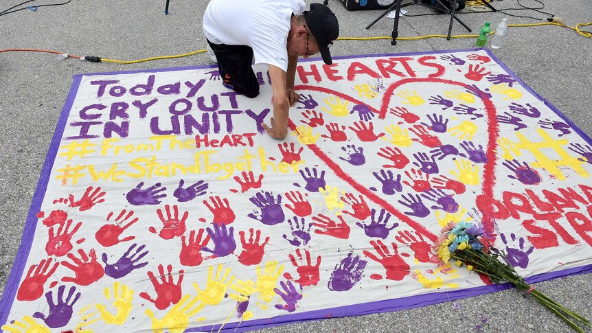A man places a hand print on a memorial of dozens of hand prints on a large sheet of paper on the ground.