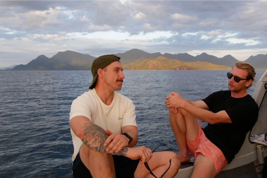 Two men relax on a boat with the ocean and a mountain range in the background.