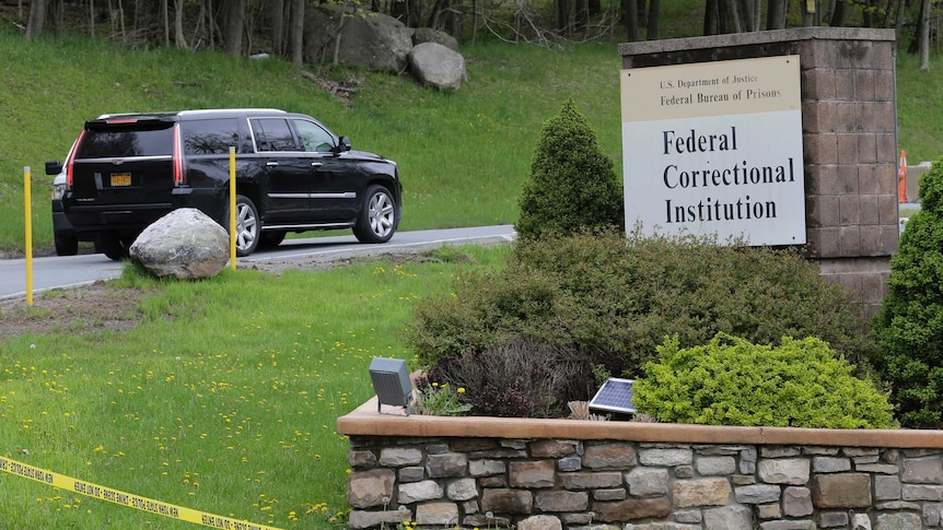 A black car drives into a driveway amid green tress with a sign out front reading 'Federal Correctional Institution'.