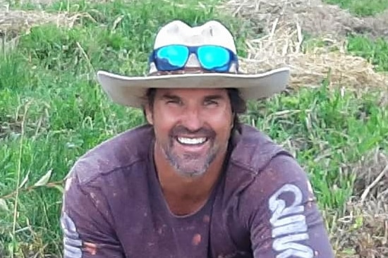 A man in a cowboy hat with sunglasses perched on the brim squats in the grass, wearing a long-sleeve purple T-shirt.