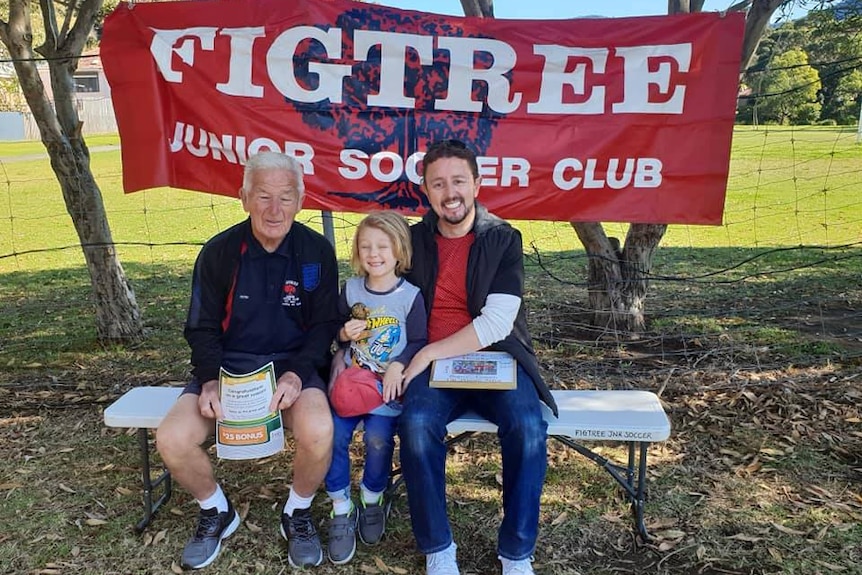 Peter, Sonny and Neil Webster sit on an outdoor seat in front of a Figtree Junior Soccer Club sign.