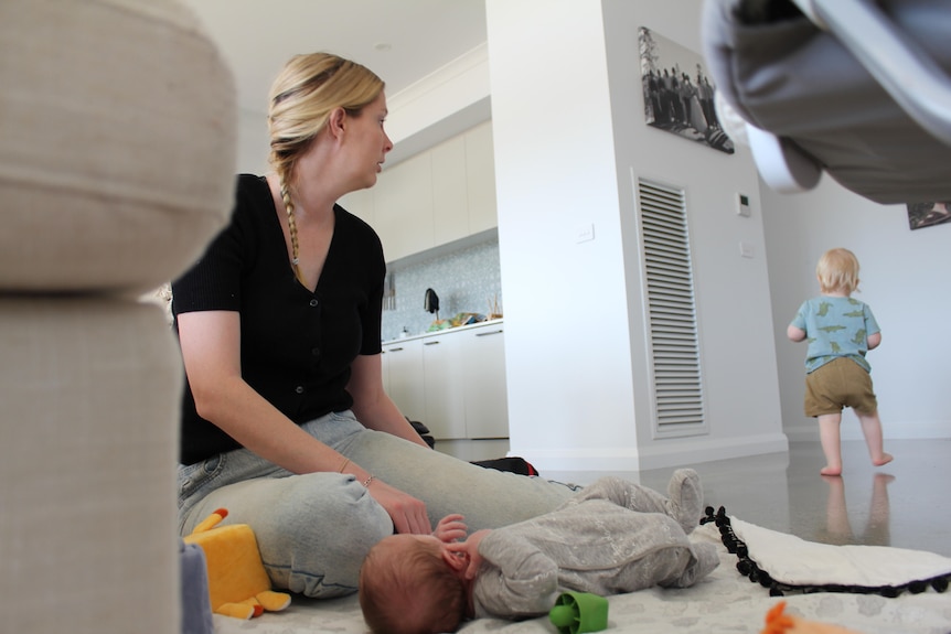 A baby is on the floor while his mother sits near him and toddler walks in the background