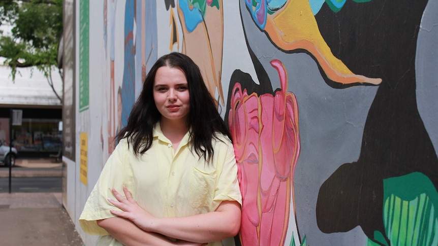 Young woman with dark straight hair stands in front of colourfully painted wall