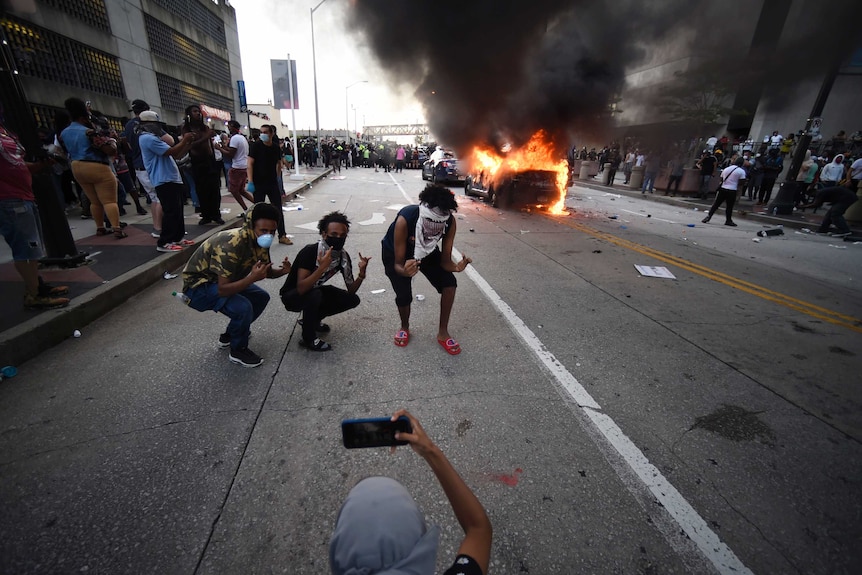 Three men signal and pose for a photo with a burning car in the background.