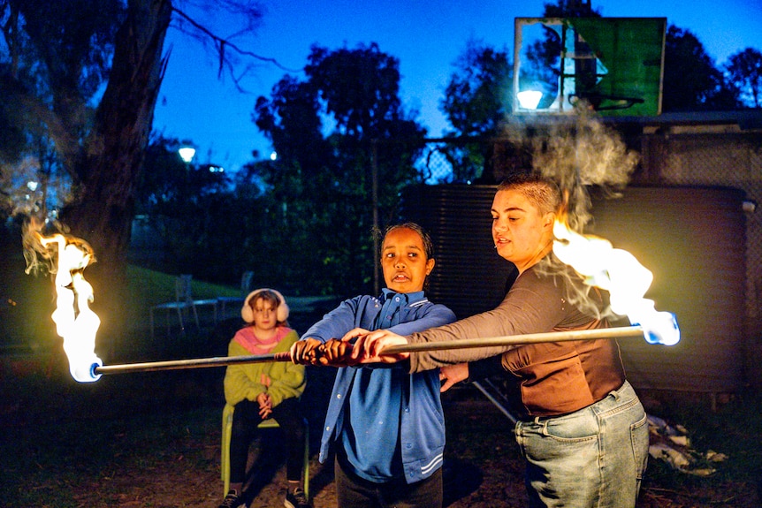 A young adult helps a young girl who is holding a stick with flames on the end. They're in a park at duskj