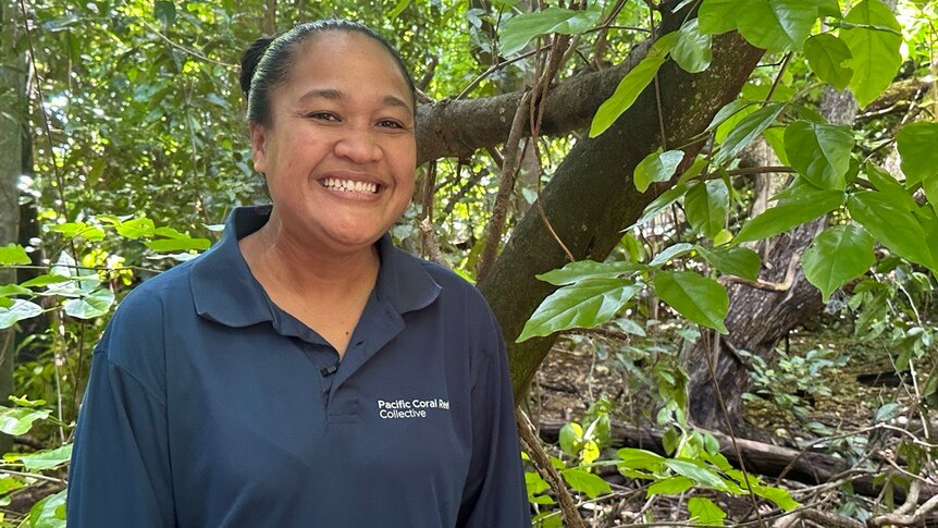 A woman smiles at the camera while standing in lush, green jungle. She's wearing a navy blue collared shirt.